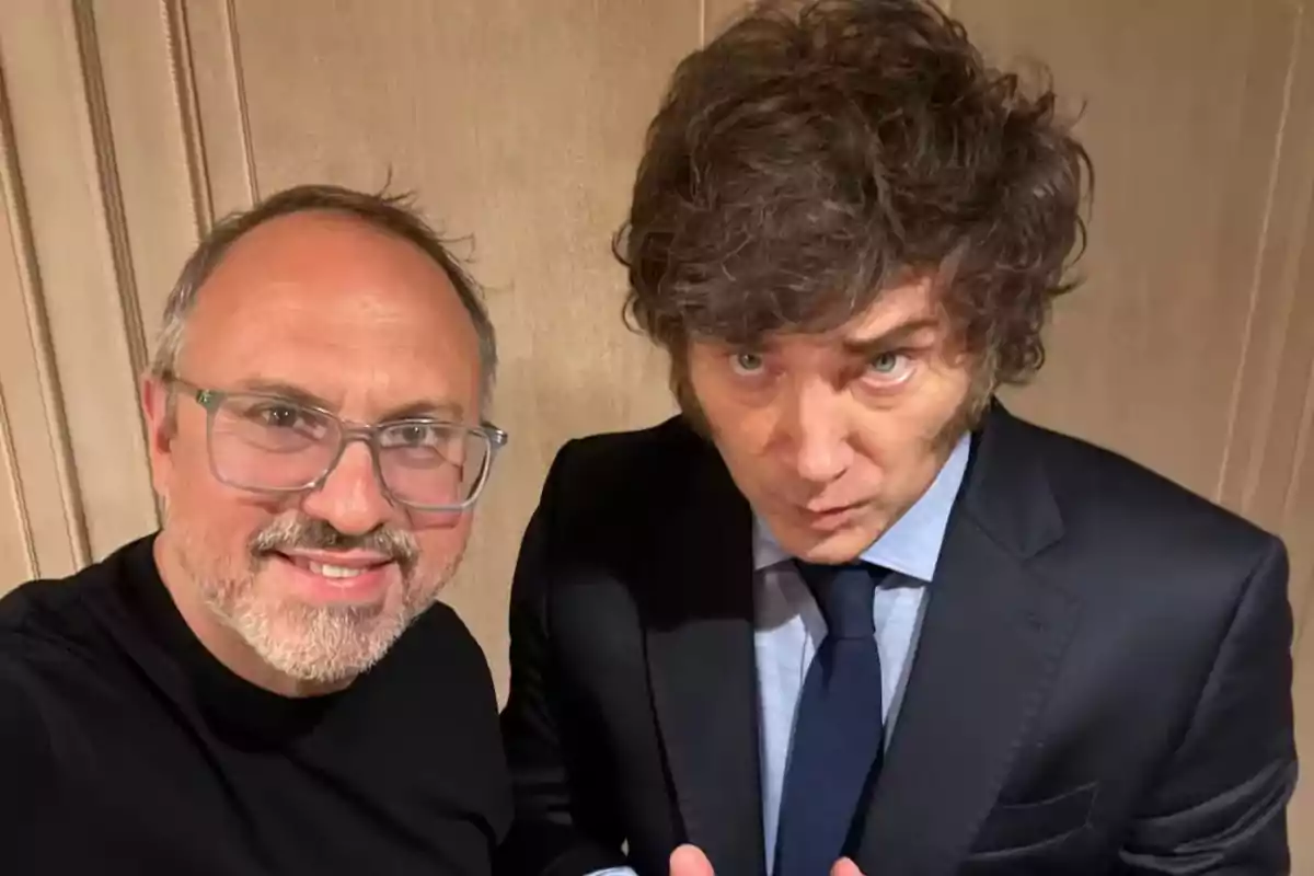 Two men pose together for a selfie, one wearing glasses and a black t-jersey, the other dressed in a dark suit and blue tie.
