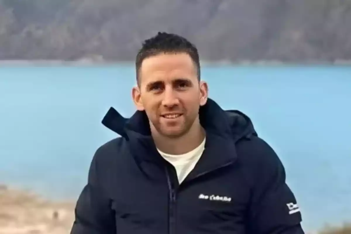 Un hombre con chaqueta oscura sonriendo frente a un lago y montañas.