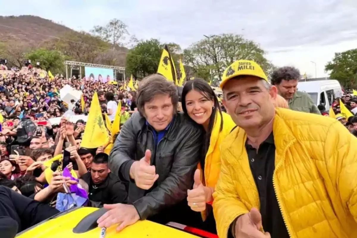 Un grupo de personas sonríe y hace el gesto de pulgar arriba en un evento al aire libre con una multitud y banderas amarillas de fondo.
