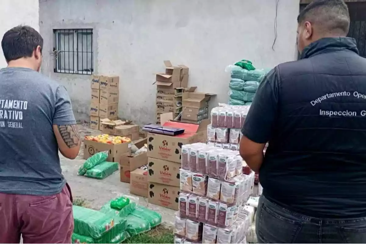 Dos personas inspeccionan un lote de cajas y productos apilados en un área exterior.