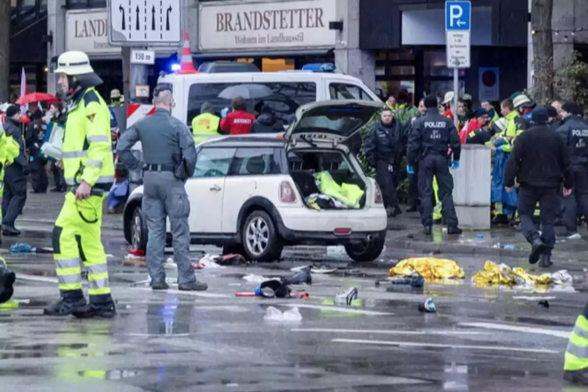 Un grupo de policías y personal de emergencia rodea un coche blanco con el maletero abierto en una calle donde hay escombros y mantas de emergencia esparcidas.