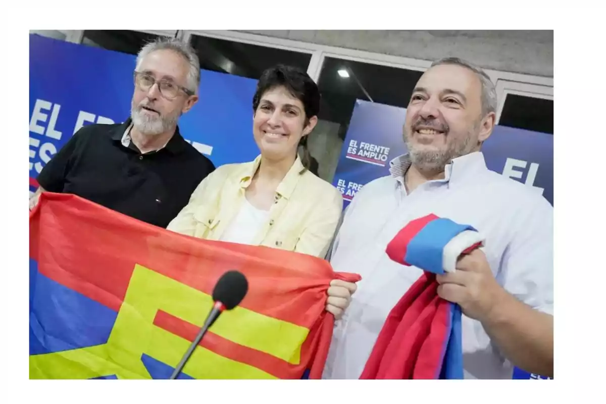 Tres personas sostienen una bandera colorida frente a un fondo azul con texto. Tres personas sostienen una bandera colorida frente a un fondo azul con texto.