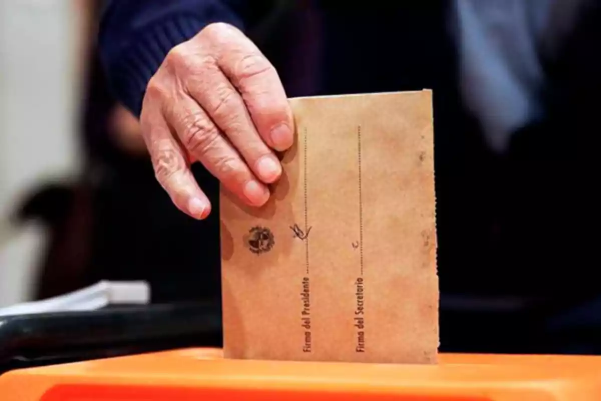 Una mano introduciendo un sobre en una urna de votación.
