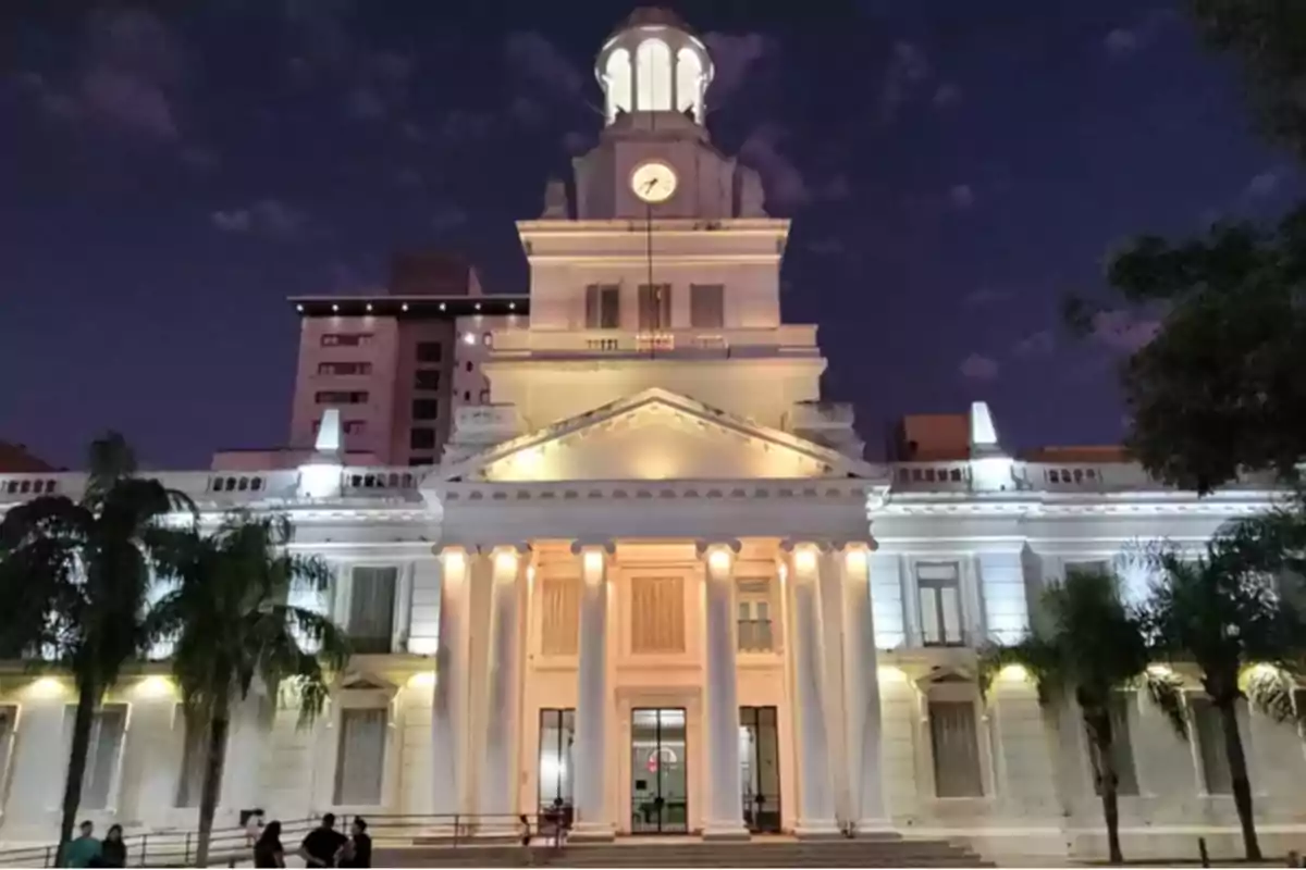 Fachada de un edificio histórico iluminado por la noche con columnas blancas y un reloj en la parte superior Fachada de un edificio histórico iluminado por la noche con columnas blancas y un reloj en la parte superior