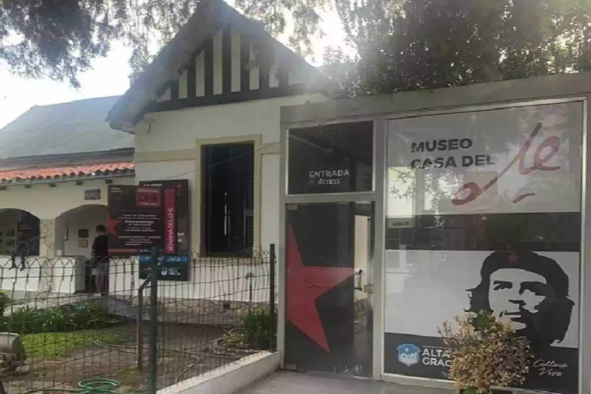 Facade of the Che House Museum with a large sign displaying the face of Ernesto Che Guevara and the main entrance to the building surrounded by vegetation and a low fence
