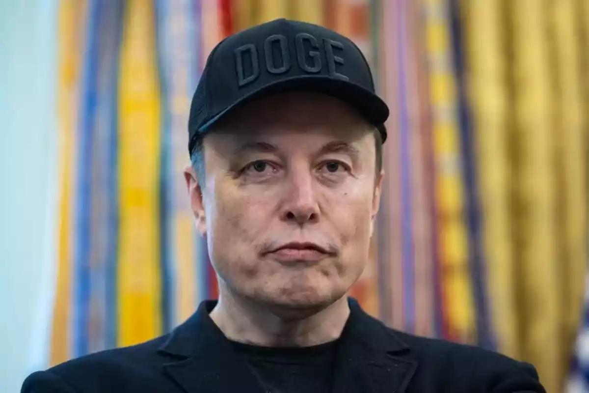 Hombre con gorra negra que tiene la palabra DOGE en relieve, fondo desenfocado con cortinas de colores. Hombre con gorra negra que tiene la palabra DOGE en relieve, fondo desenfocado con cortinas de colores.