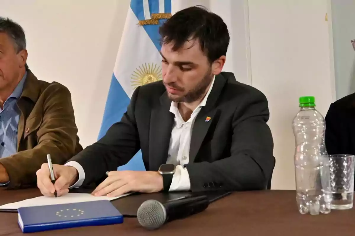 Un hombre firmando un documento en una mesa con una bandera de Argentina de fondo.