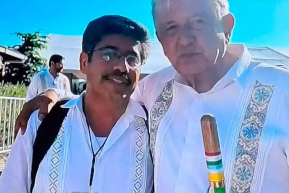 Two men in white guayaberas pose together outdoors on a sunny day, one of them holding a decorated cane, and both smiling at the camera.