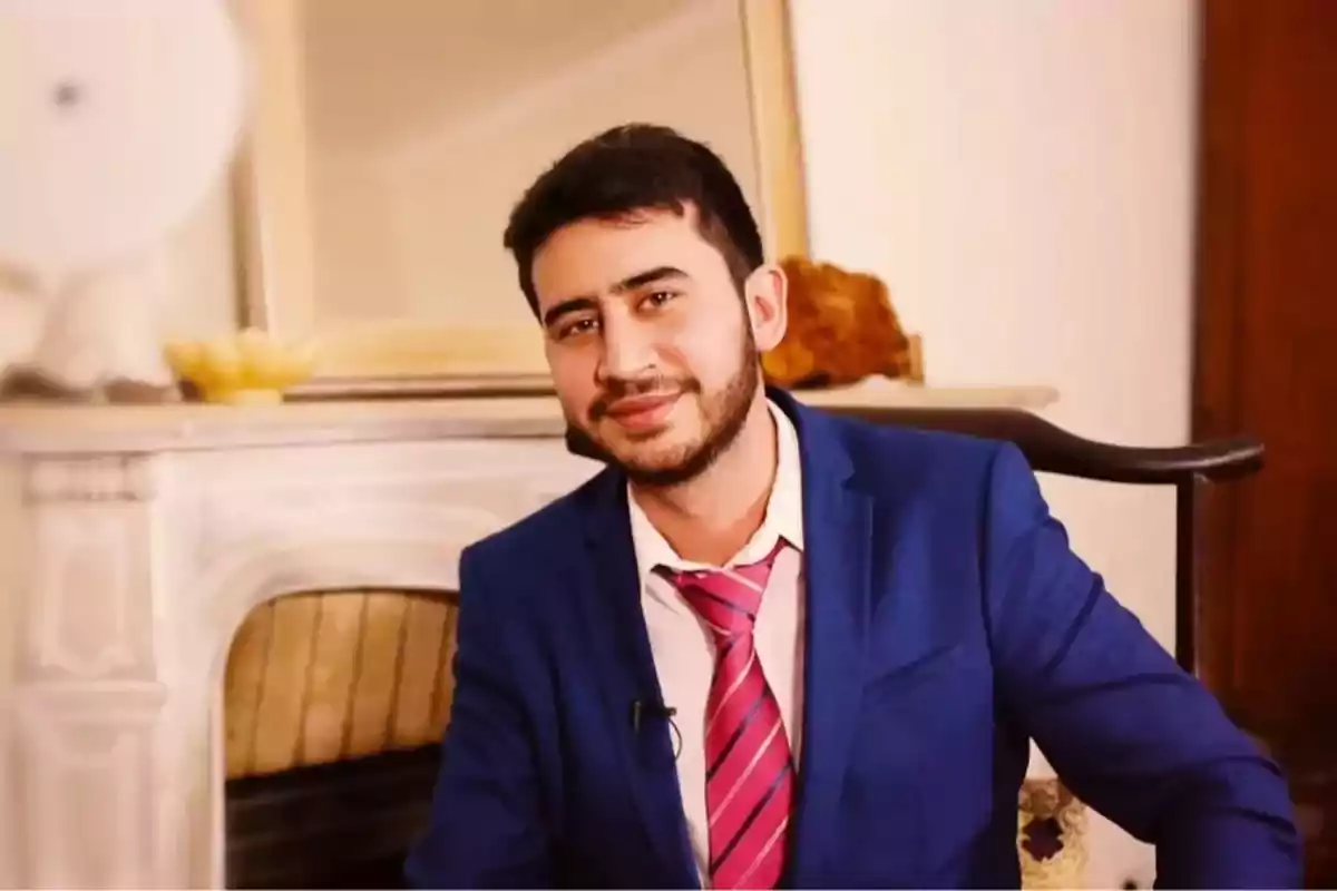 Hombre joven con barba y cabello oscuro viste traje azul y corbata rosa mientras sonríe sentado en una silla frente a una chimenea blanca