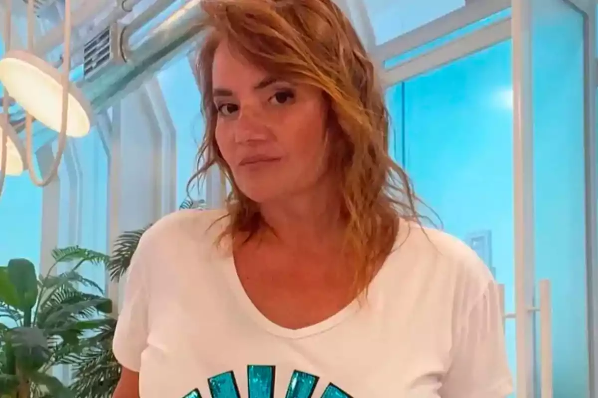 Light brown, wavy-haired woman wearing a white t-jersey posing in a modern interior with plants and large windows in the background Light brown, wavy-haired woman wearing a white t-jersey posing in a modern interior with plants and large windows in the background
