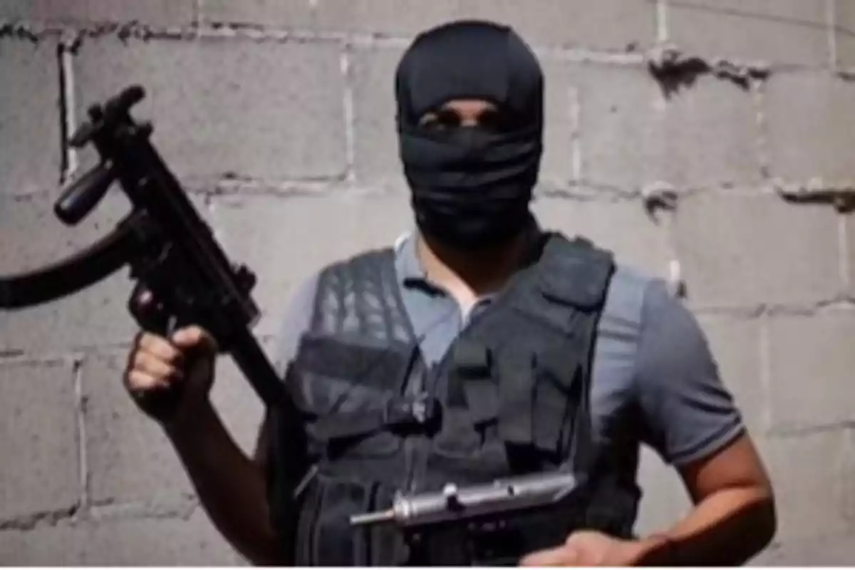 A masked man in a tactical vest holds a gun in front of a concrete block wall.