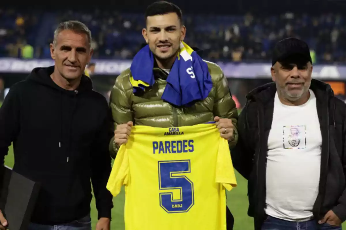 Tres hombres posan en un estadio de fútbol mientras uno de ellos sostiene una camiseta amarilla con el nombre Paredes y el número 5 Tres hombres posan en un estadio de fútbol mientras uno de ellos sostiene una camiseta amarilla con el nombre Paredes y el número 5