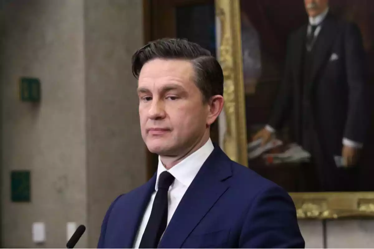 A man in a dark blue suit and black tie is standing in front of a microphone with a framed portrait in the background. A man in a dark blue suit and black tie is standing in front of a microphone with a framed portrait in the background.