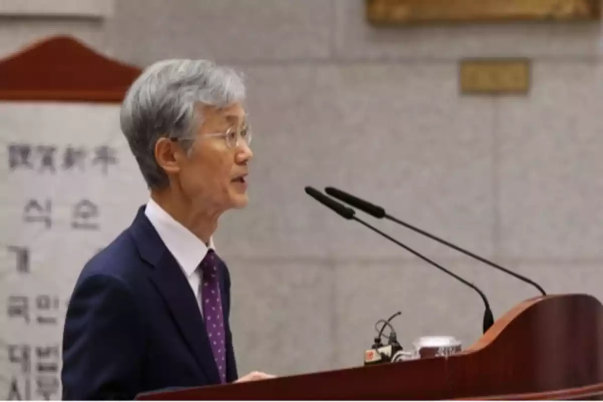 Gray-haired man with glasses speaking at a podium with microphones in a formal setting
