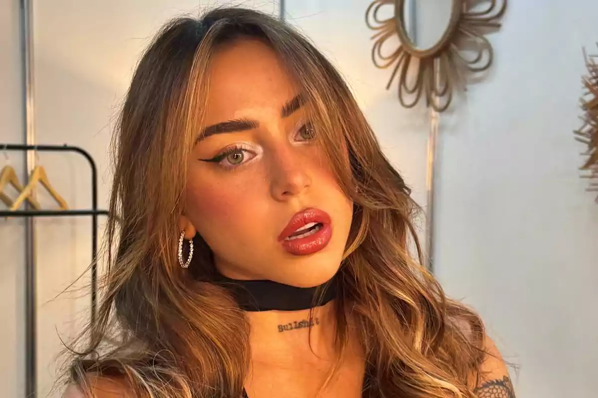 Una persona con cabello largo y maquillaje llamativo, usando aretes y un collar negro, en un entorno interior con espejos decorativos.