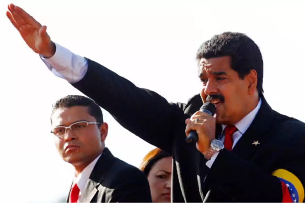 Un hombre con traje oscuro y corbata roja sostiene un micrófono mientras levanta el brazo, acompañado de otra persona con gafas y traje similar. Un hombre con traje oscuro y corbata roja sostiene un micrófono mientras levanta el brazo, acompañado de otra persona con gafas y traje similar.