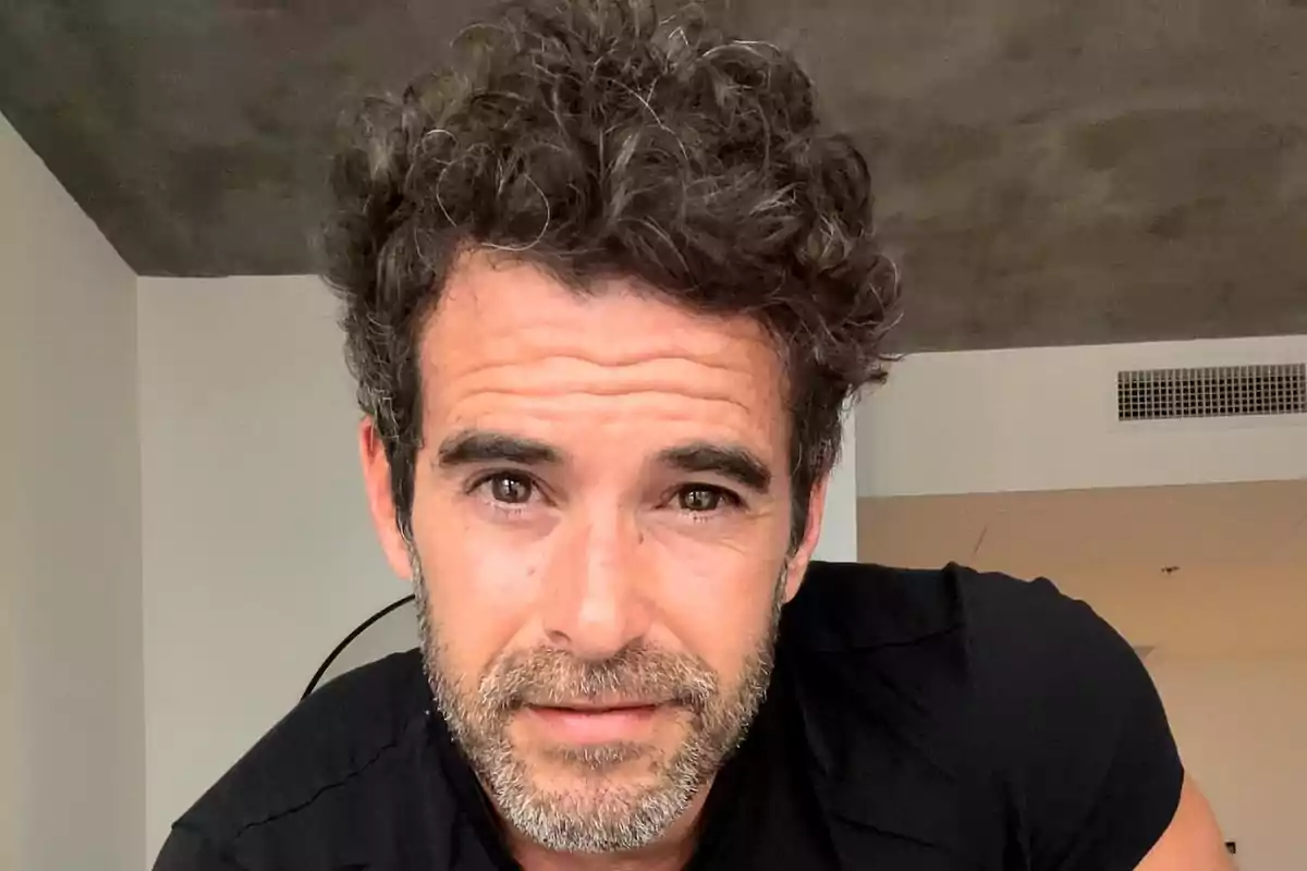 Hombre de cabello rizado y barba corta mirando a la cámara en una habitación iluminada