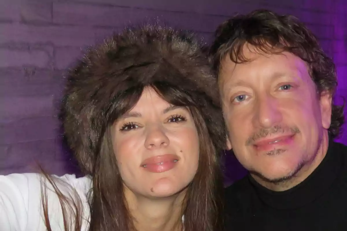 Una mujer con gorro de peluche marrón y un hombre con suéter negro posan juntos y sonríen frente a una pared iluminada con luz violeta