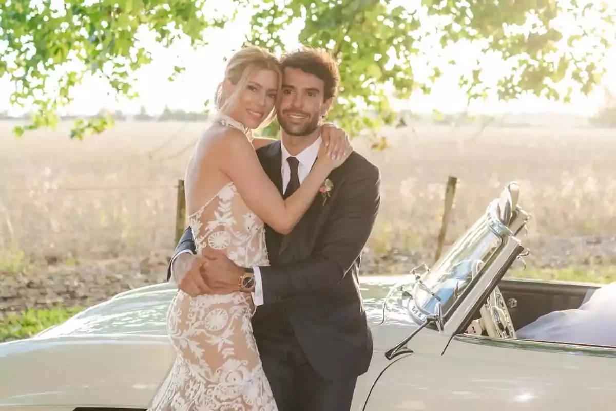 Pareja de recién casados abrazados junto a un auto clásico en un entorno campestre y soleado Pareja de recién casados abrazados junto a un auto clásico en un entorno campestre y soleado