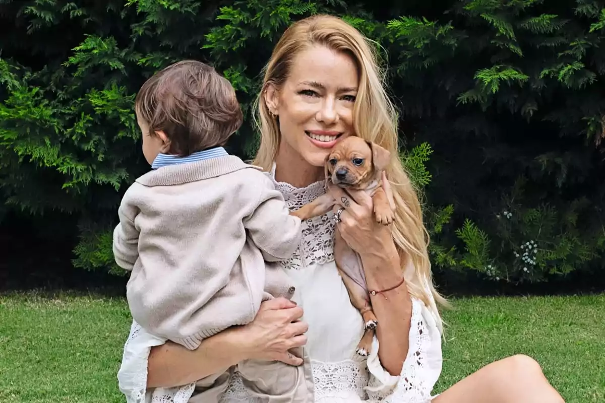 Una mujer sonriente sostiene a un cachorro y a un niño pequeño en un jardín con arbustos verdes de fondo.