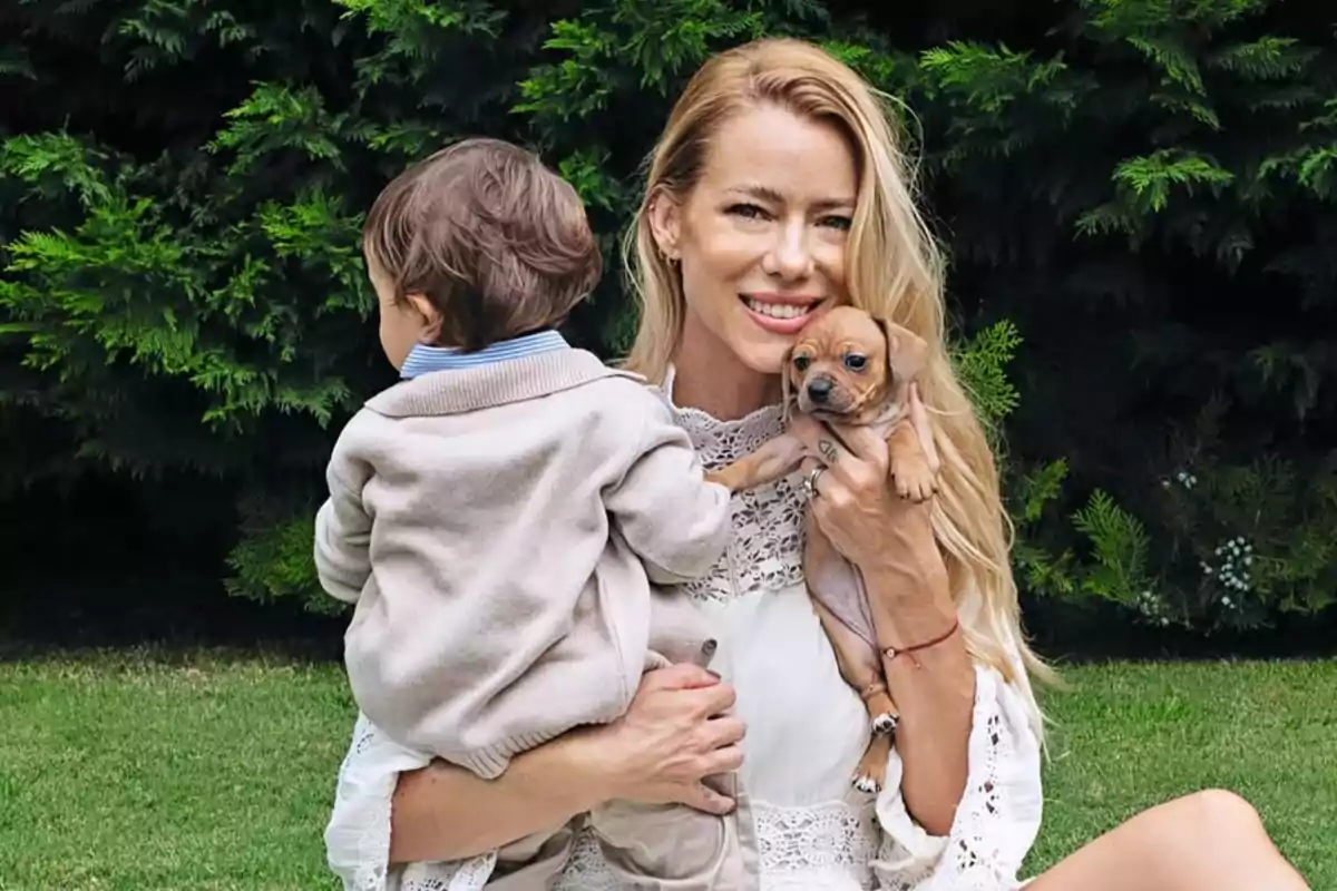Smiling woman sitting on the grass holding a baby and a small puppy in front of some green bushes Smiling woman sitting on the grass holding a baby and a small puppy in front of some green bushes