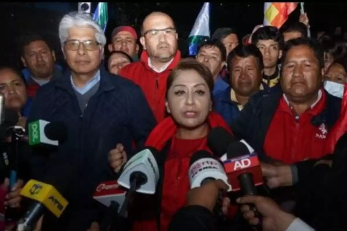 A woman surrounded by microphones gives statements to the press while several people accompany her in the background. A woman surrounded by microphones gives statements to the press while several people accompany her in the background.