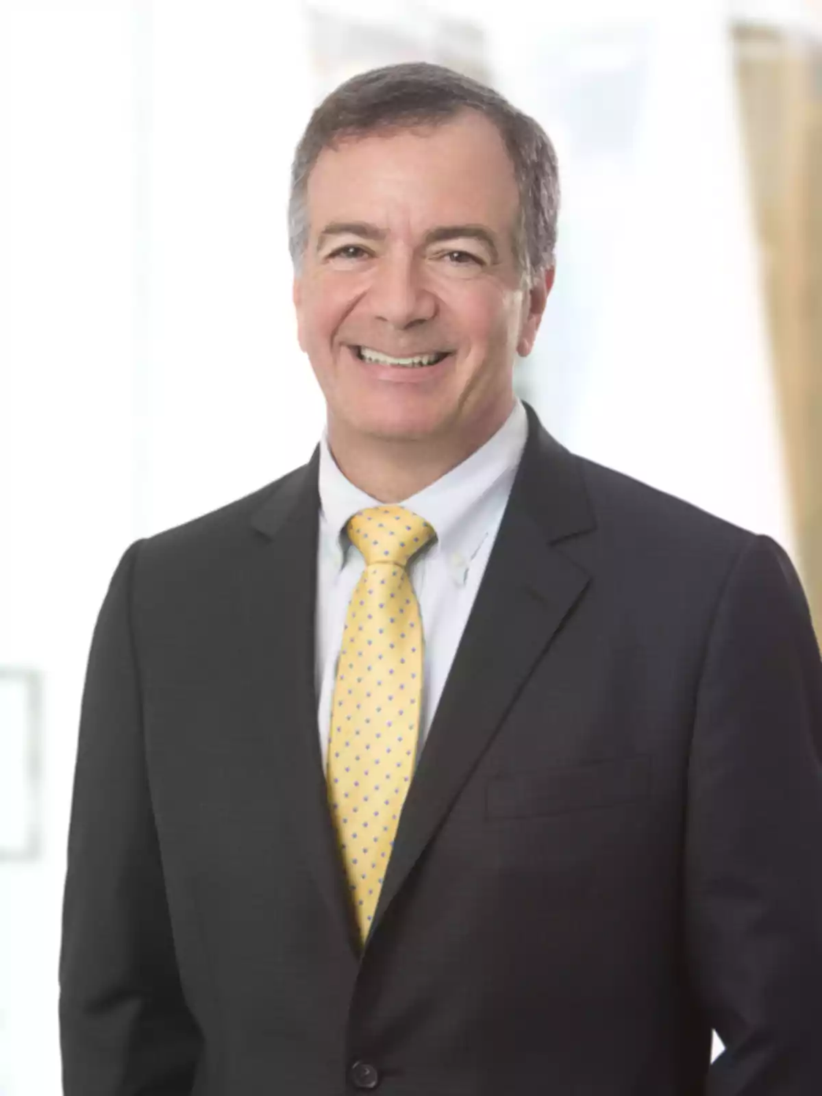 A man smiling in a dark suit and a yellow tie with blue dots on a light background. A man smiling in a dark suit and a yellow tie with blue dots on a light background.