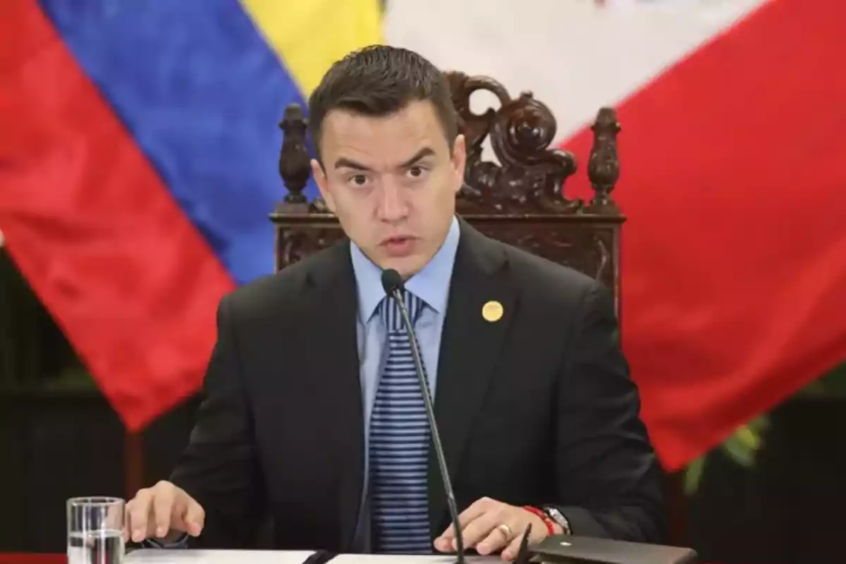 Un hombre en traje oscuro habla en un micrófono frente a banderas de colores rojo, amarillo y azul.