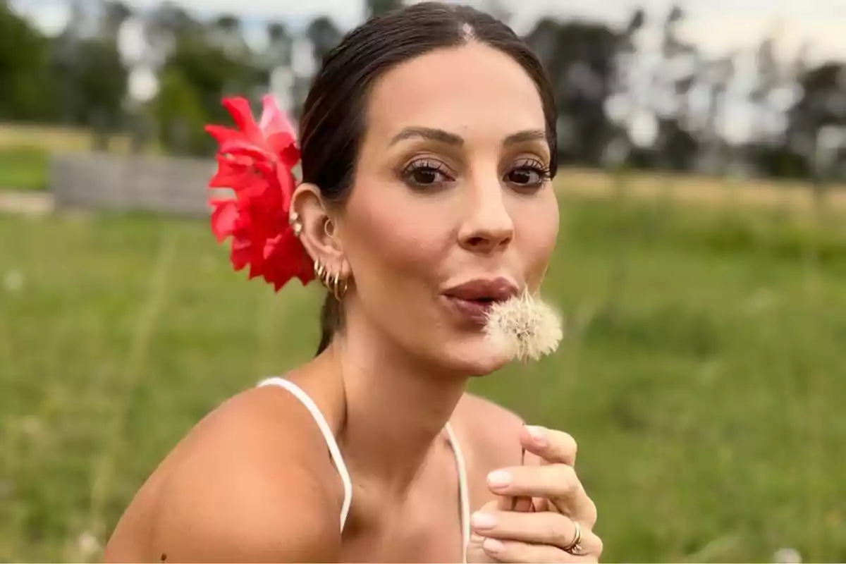 Mujer con una flor roja en el cabello soplando un diente de león en un campo verde.