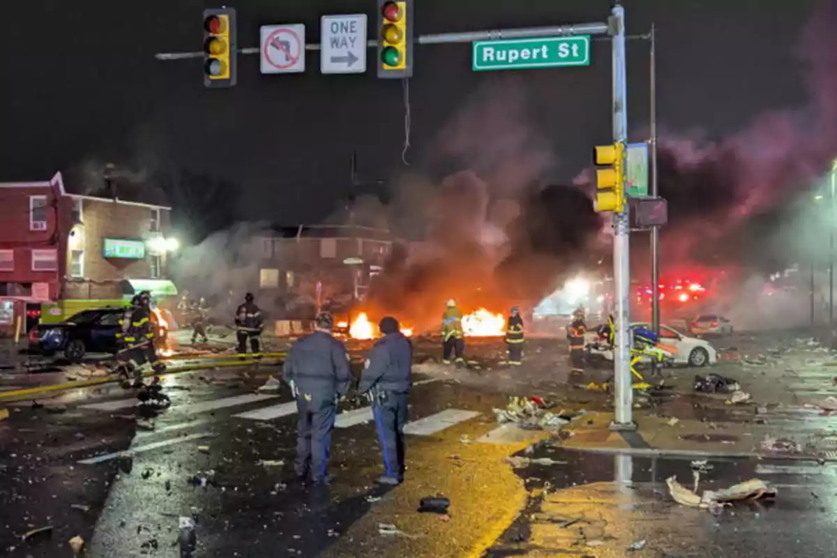 Una escena nocturna muestra a bomberos y policías respondiendo a un incendio en una intersección urbana con escombros esparcidos por el suelo y señales de tráfico visibles.