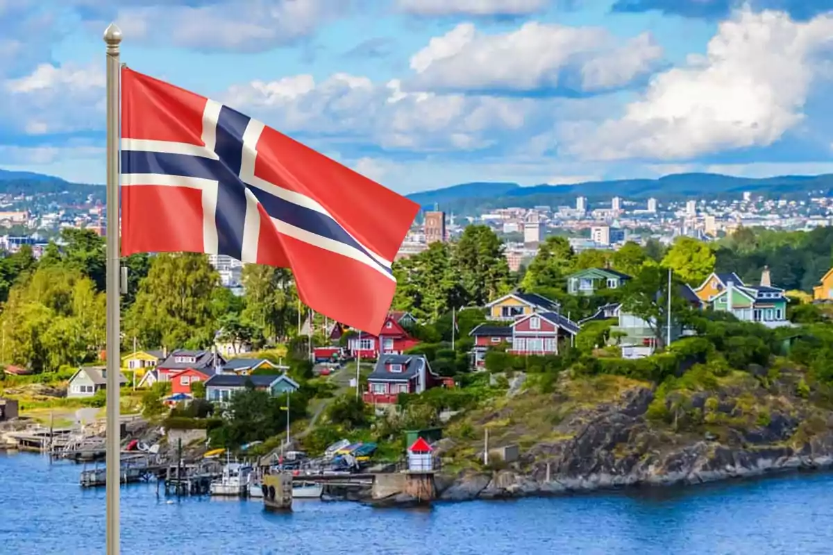 Bandera de Noruega ondeando frente a un paisaje urbano con casas coloridas y vegetación.