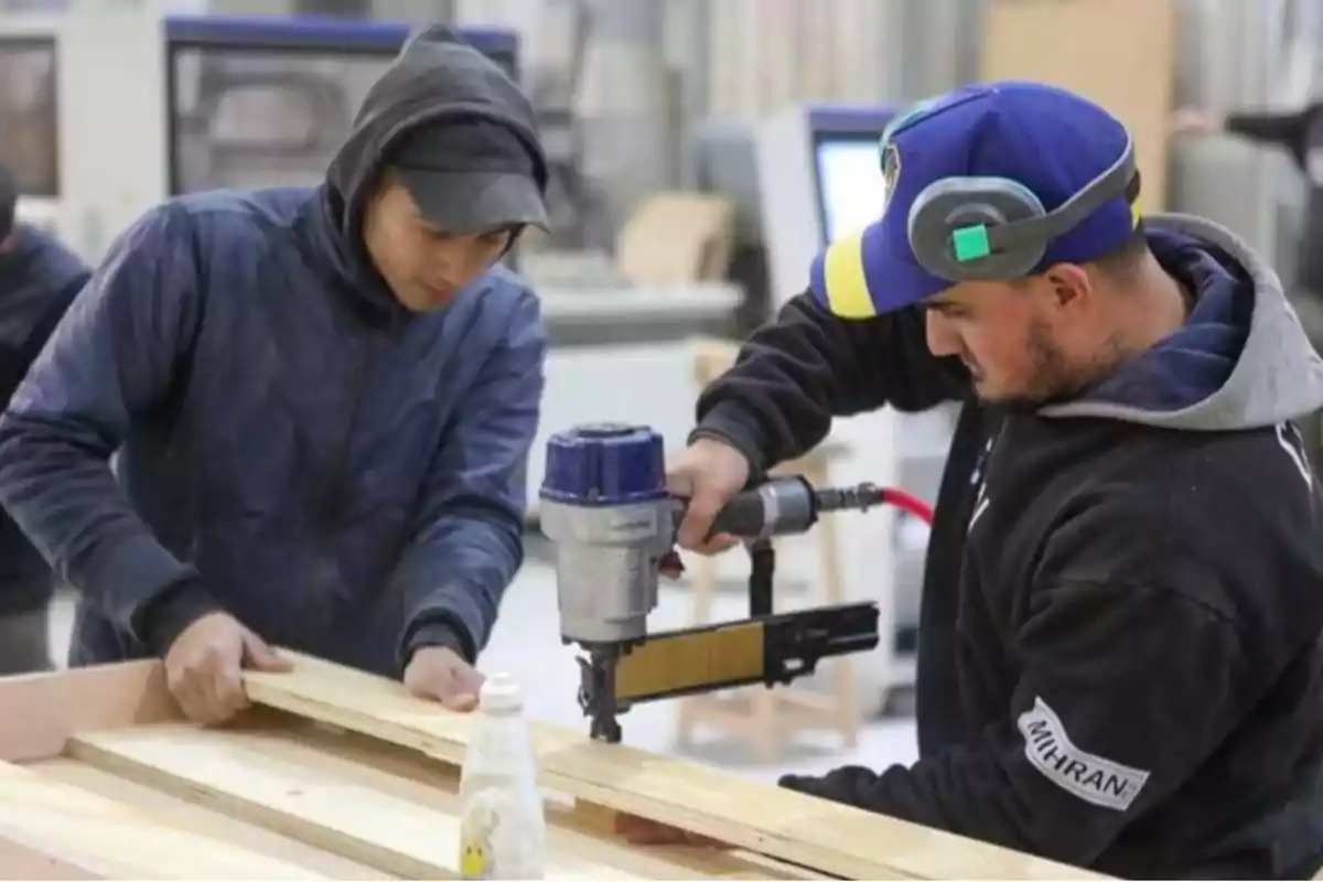 Dos personas trabajando en un taller de carpintería, una de ellas usando una pistola de clavos para ensamblar piezas de madera.