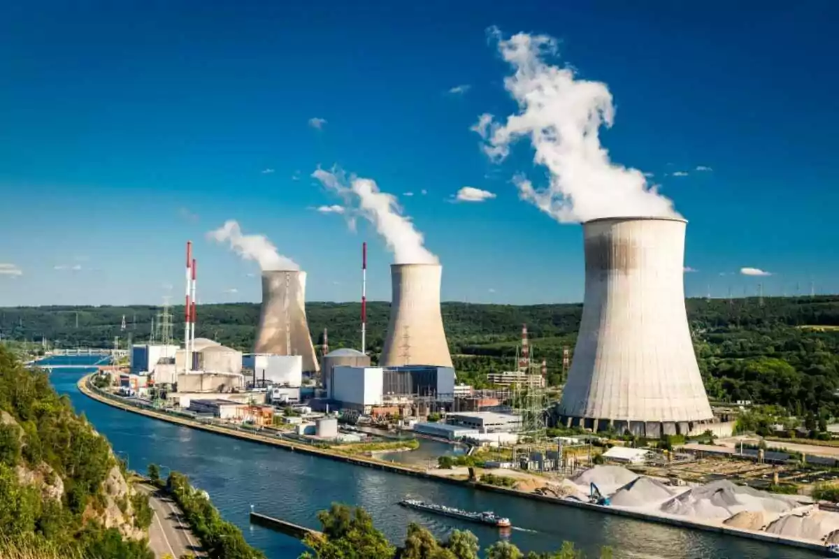 Planta de energía nuclear con torres de enfriamiento emitiendo vapor cerca de un río en un entorno verde y montañoso bajo un cielo azul.