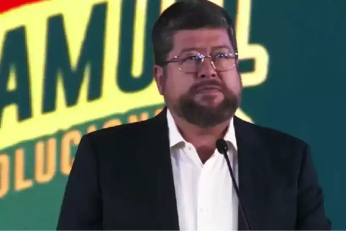 Un hombre con barba y gafas está hablando frente a un micrófono con un fondo colorido que tiene letras grandes. Un hombre con barba y gafas está hablando frente a un micrófono con un fondo colorido que tiene letras grandes.