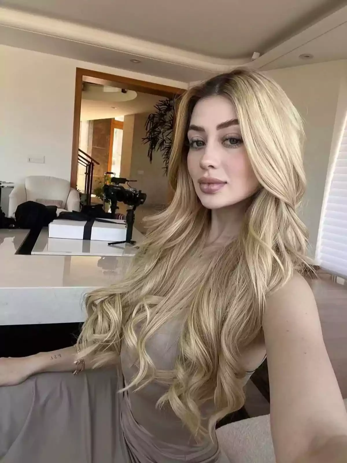 Una persona con cabello rubio largo y ondulado toma una selfie en una habitación moderna con muebles claros y una cámara en el fondo. Una persona con cabello rubio largo y ondulado toma una selfie en una habitación moderna con muebles claros y una cámara en el fondo.