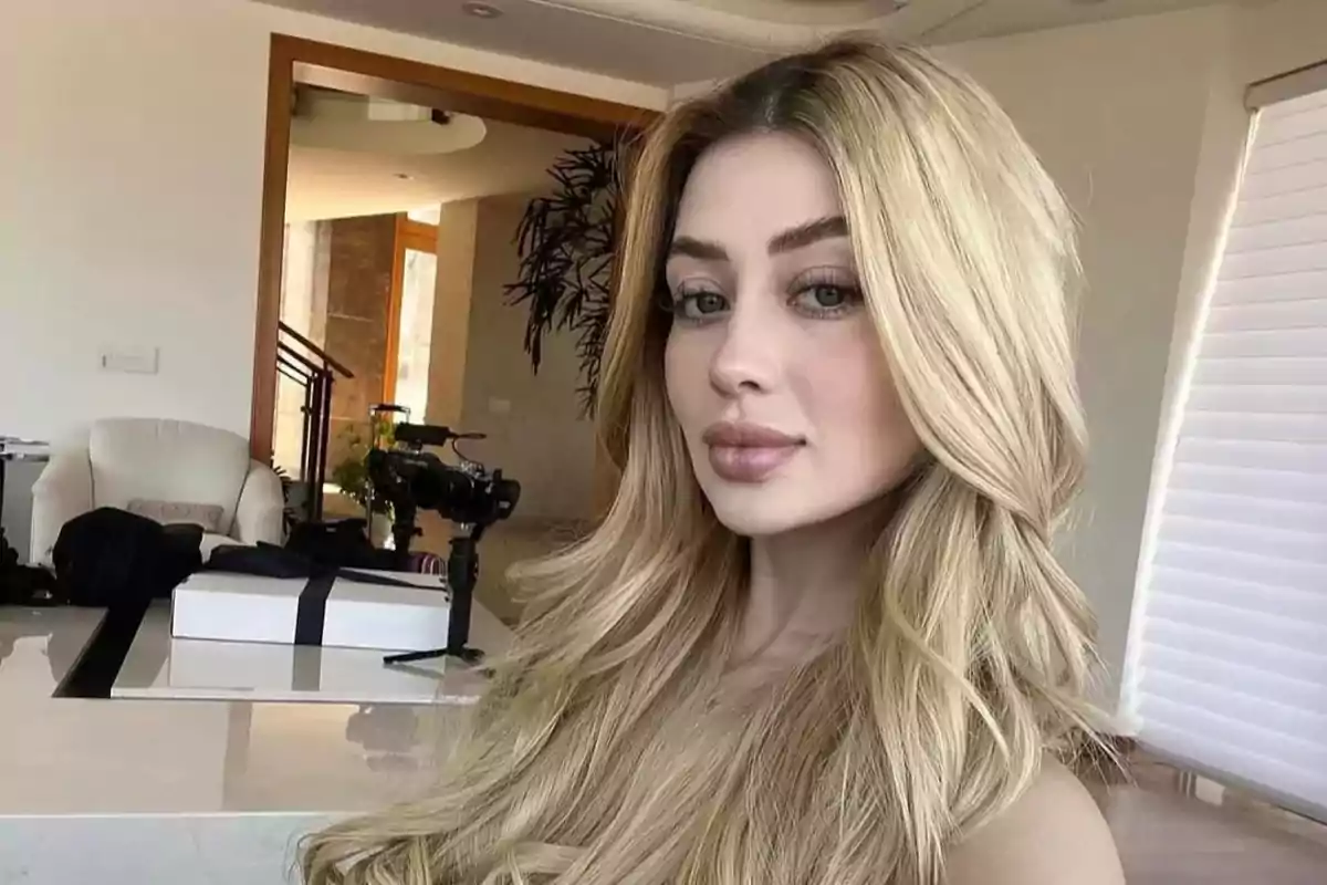Una persona con cabello rubio largo y ondulado posa en una habitación moderna con muebles claros y una cámara en el fondo.