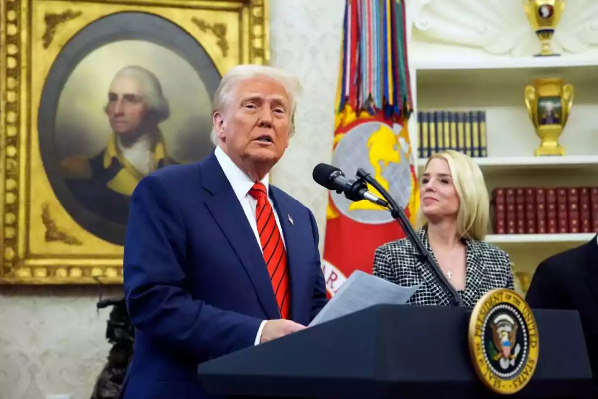Blond-haired man in a blue suit and red tie speaking at a presidential podium, accompanied by a blonde woman in an office decorated with a portrait and flags.