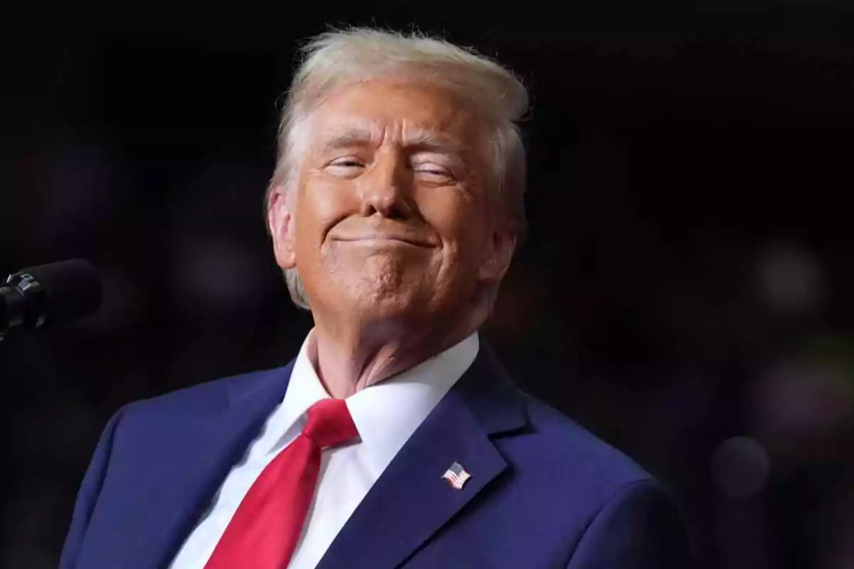 Hombre de cabello rubio con traje azul y corbata roja sonriendo frente a un micrófono