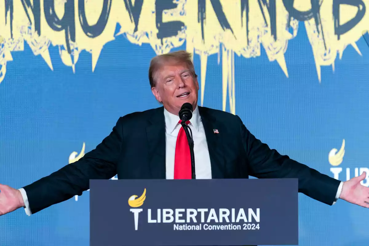 Un hombre de traje oscuro y corbata roja habla en un podio con el logo y texto de la Convención Nacional Libertaria 2024 sobre un fondo azul