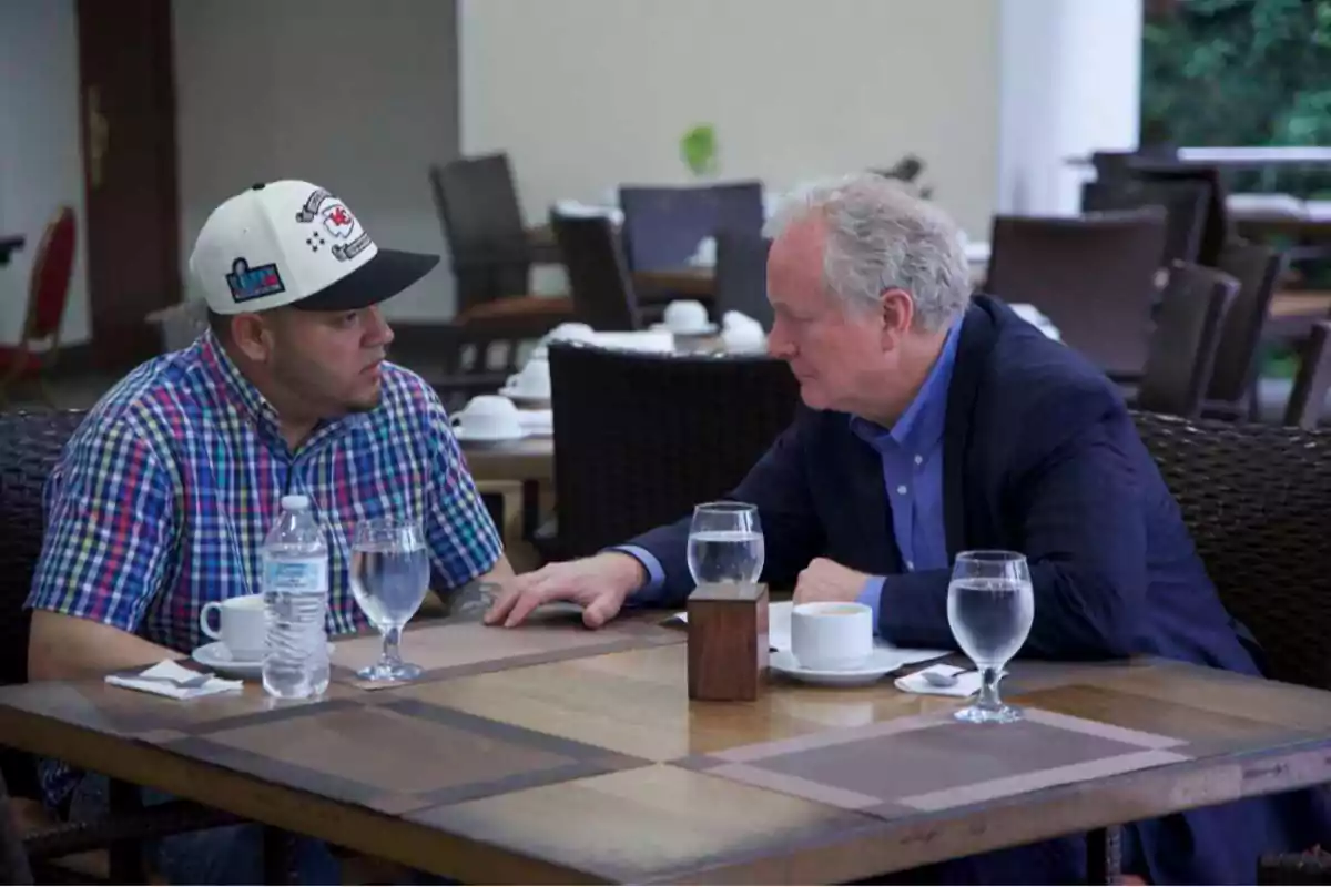 Two men sitting at a restaurant table are having a serious conversation while they have drinks and coffee cups in front of them.
