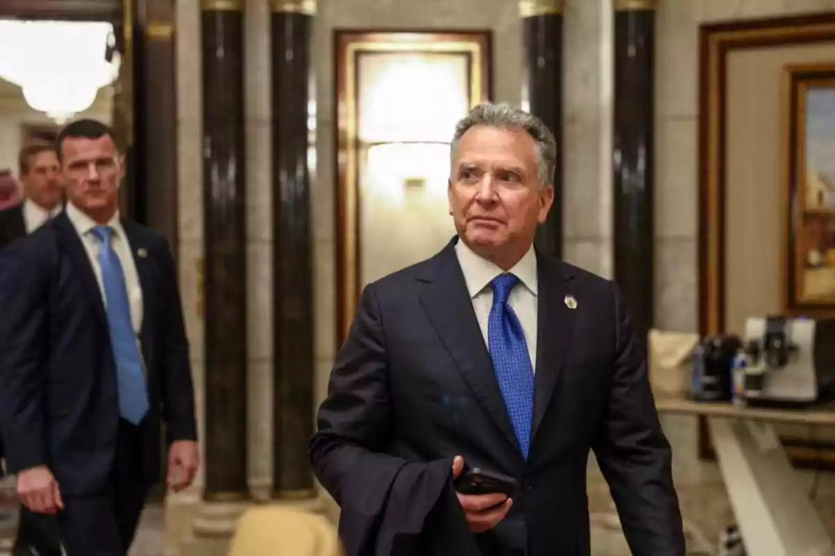 Gray-haired man in a dark suit and blue tie walking in an elegant hall accompanied by other people in the background Gray-haired man in a dark suit and blue tie walking in an elegant hall accompanied by other people in the background