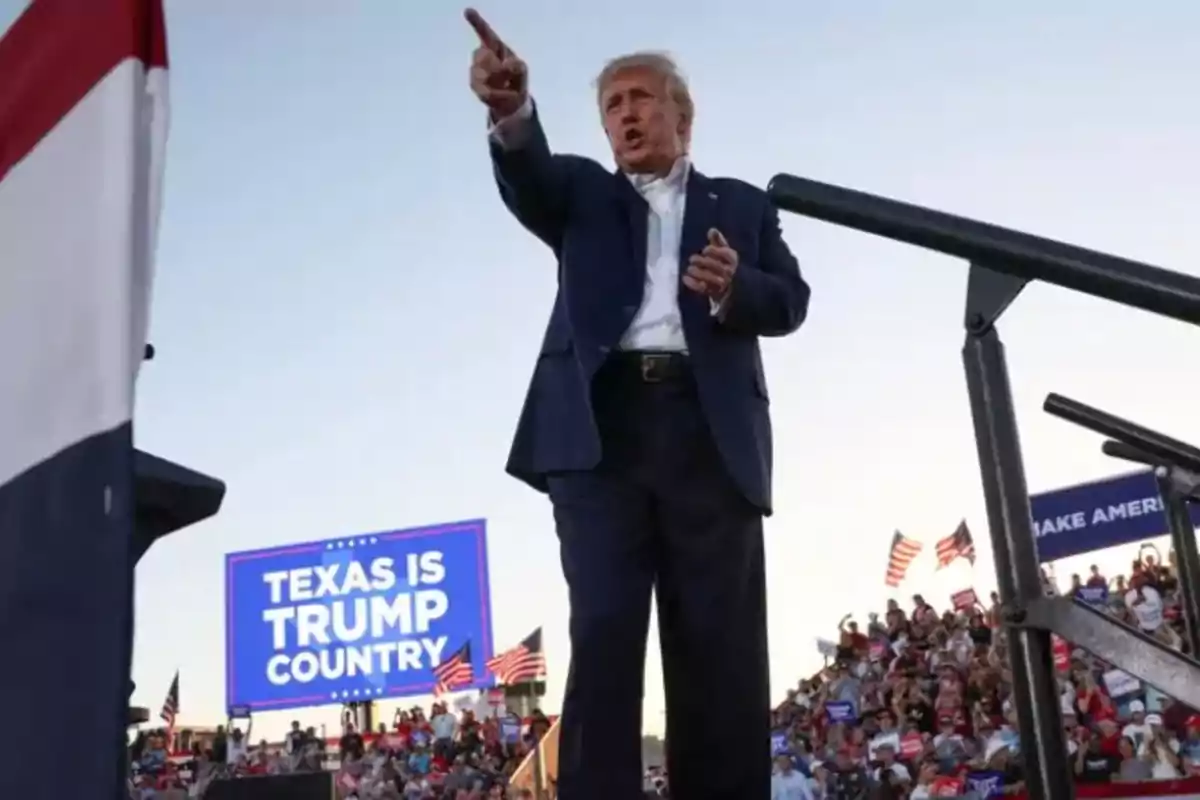 Hombre de traje azul señalando hacia el frente en un mitin político al aire libre con una multitud y un cartel que dice Texas is Trump Country de fondo