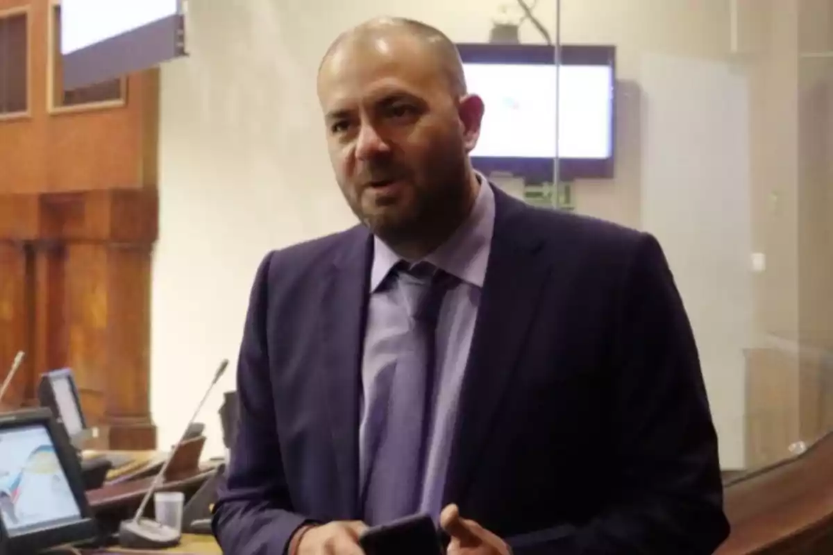 Un hombre con traje oscuro y corbata lila sostiene un teléfono en una sala con pantallas y escritorios. Un hombre con traje oscuro y corbata lila sostiene un teléfono en una sala con pantallas y escritorios.