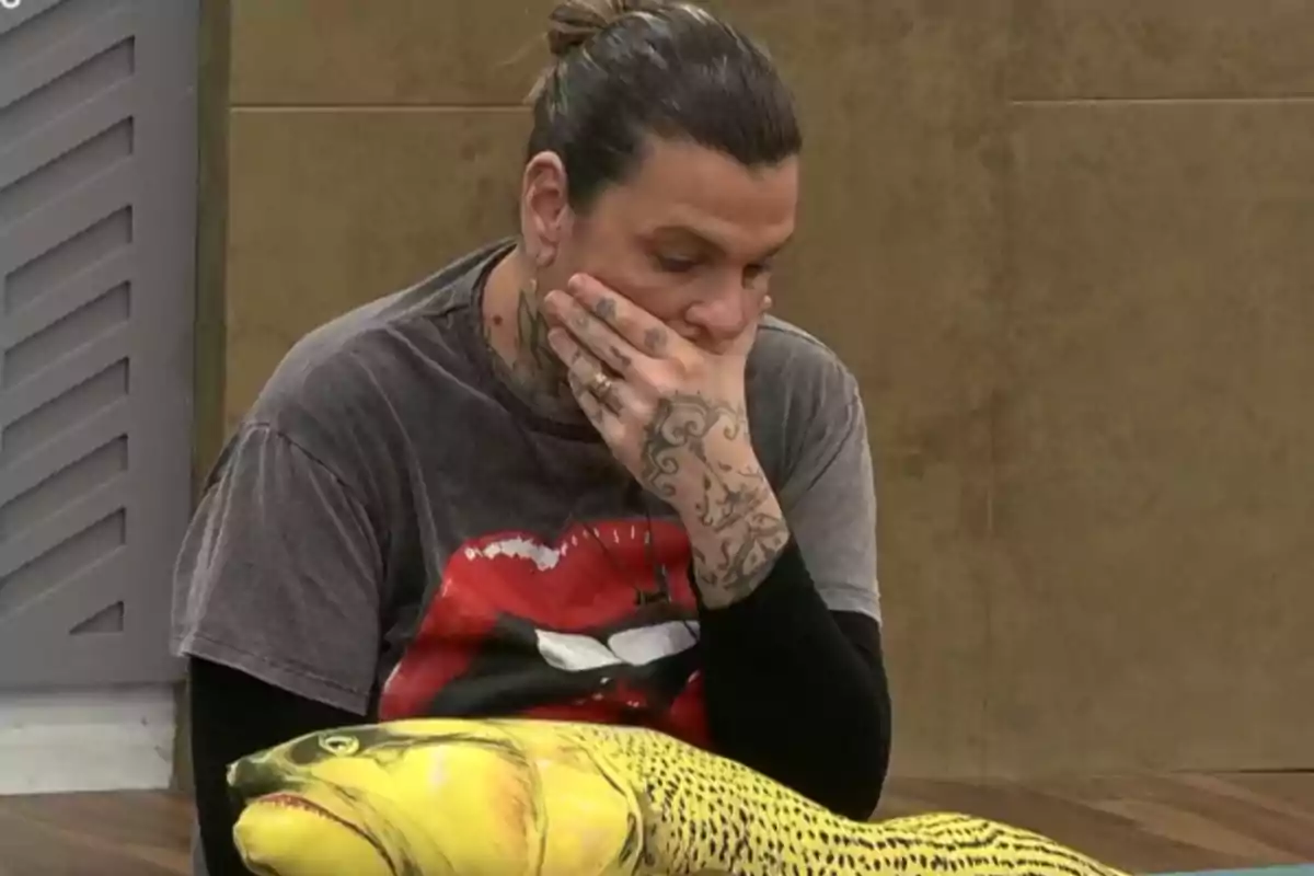 A man with tattoos on his hands and neck is sitting thoughtfully with his hand on his chin and a yellow fish-shaped cushion on his lap. A man with tattoos on his hands and neck is sitting thoughtfully with his hand on his chin and a yellow fish-shaped cushion on his lap.