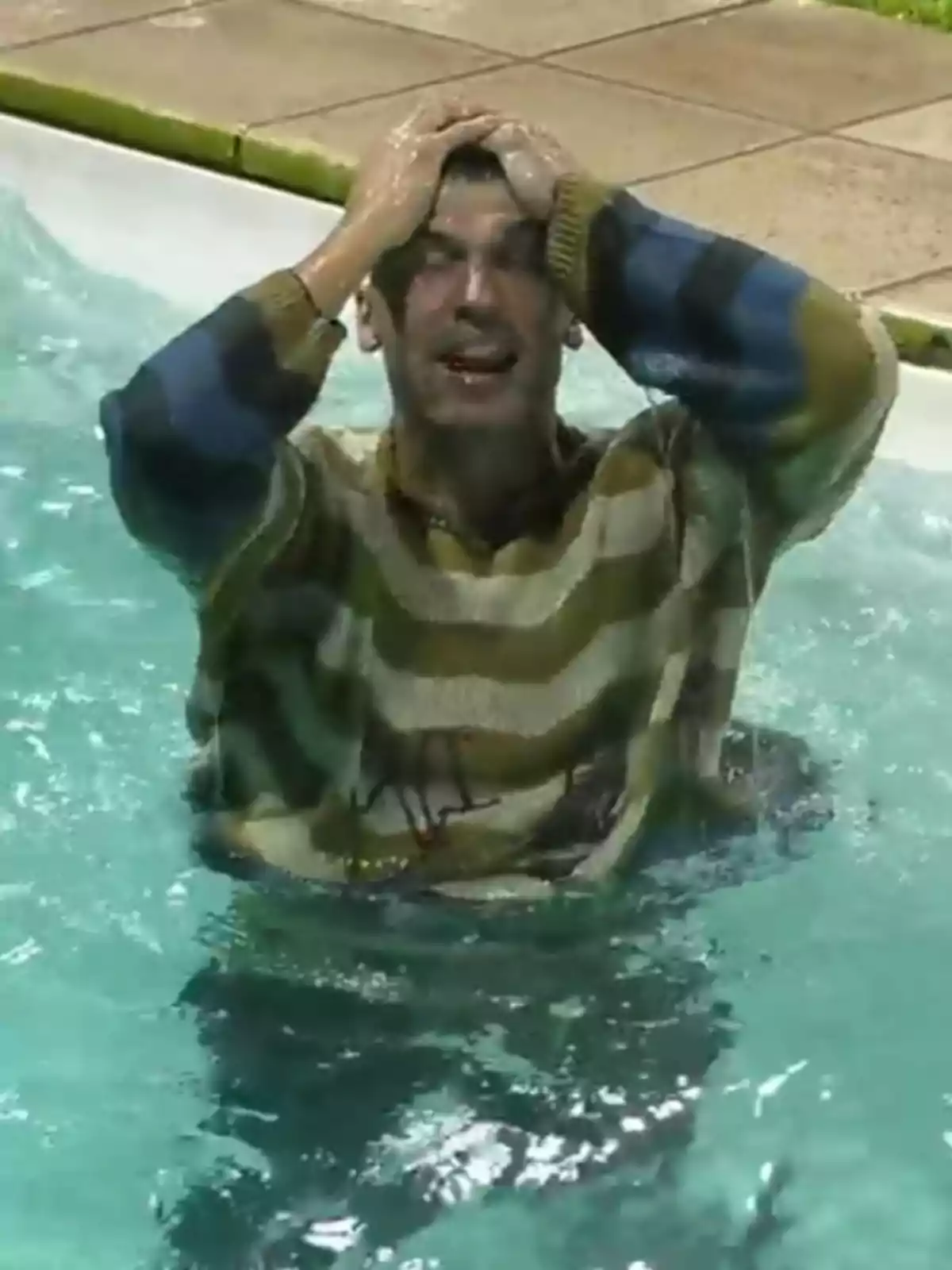 Hombre con suéter de rayas dentro de una piscina sosteniéndose la cabeza con ambas manos y expresión de sorpresa Hombre con suéter de rayas dentro de una piscina sosteniéndose la cabeza con ambas manos y expresión de sorpresa