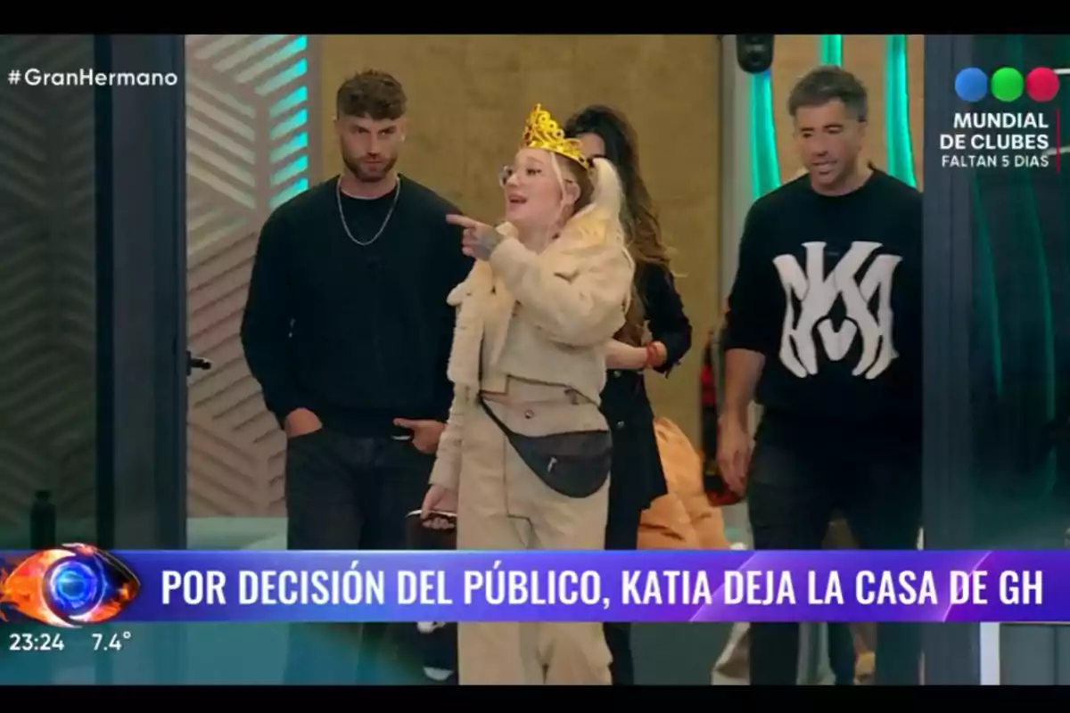 Several reality show participants are standing in a room while a woman wearing a crown and light-colored clothes points forward; at the bottom, there is an announcement about Katia leaving the Big Brother house. Several reality show participants are standing in a room while a woman wearing a crown and light-colored clothes points forward; at the bottom, there is an announcement about Katia leaving the Big Brother house.