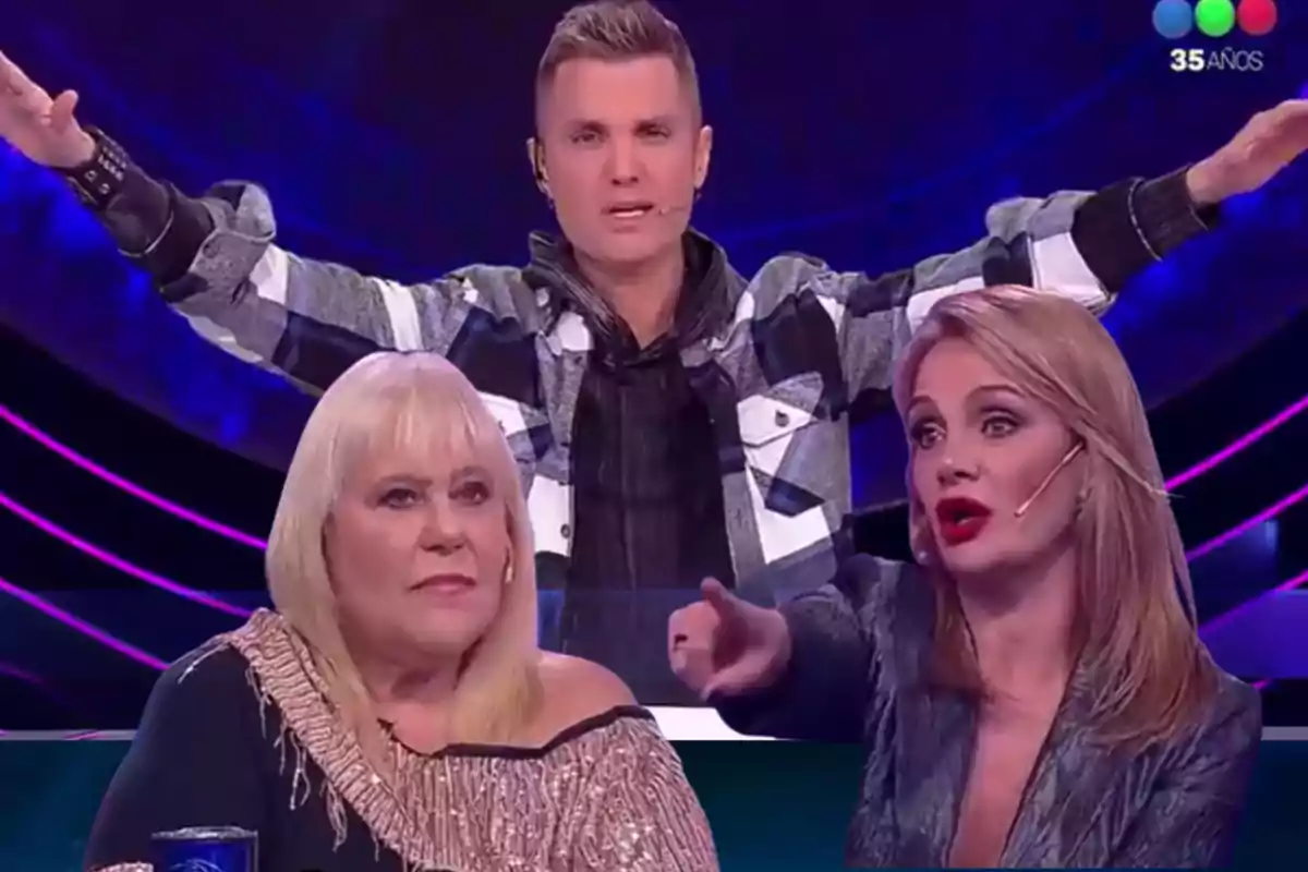 Tres personas en un set de televisión con fondo azul y luces, una mujer rubia mayor sentada, una mujer rubia joven señalando y un hombre de pie con los brazos extendidos.