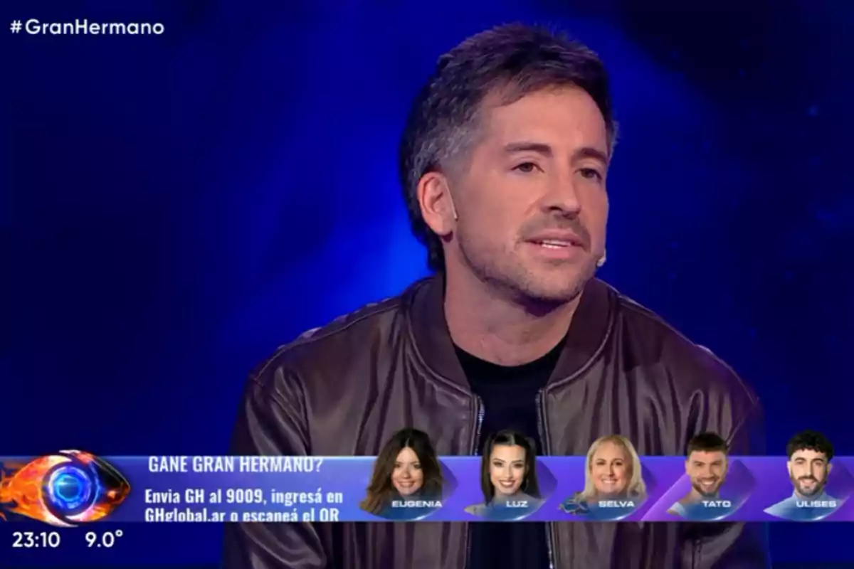 Un hombre con chaqueta marrón está en un set de televisión con fondo azul, en la pantalla aparecen fotos de cinco participantes y un gráfico de Gran Hermano junto a información para votar. Un hombre con chaqueta marrón está en un set de televisión con fondo azul, en la pantalla aparecen fotos de cinco participantes y un gráfico de Gran Hermano junto a información para votar.