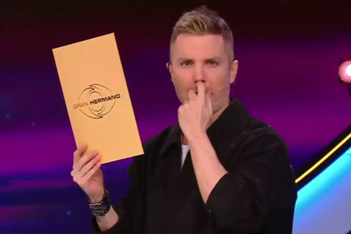 A host holds a yellow envelope with a TV show logo while bringing his hand to his mouth on a lit stage.