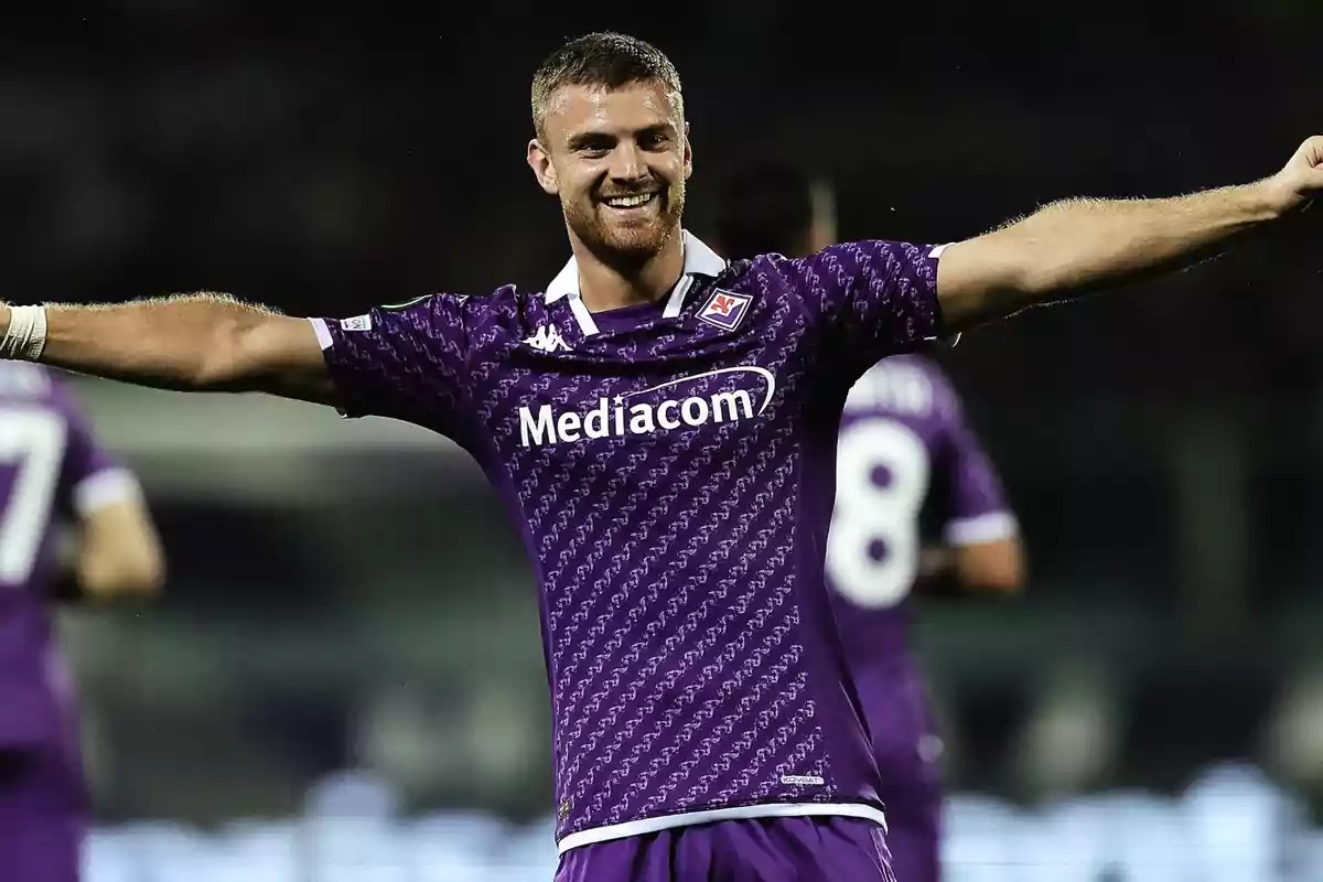 A player in a purple uniform celebrates with arms outstretched on a playing field. A player in a purple uniform celebrates with arms outstretched on a playing field.